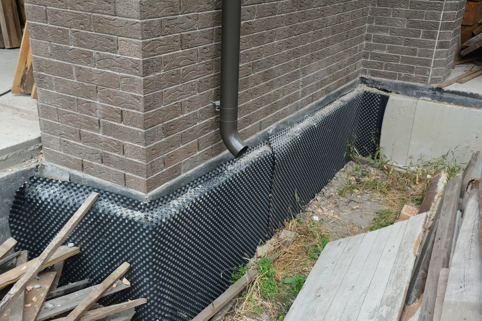 Downpipe and drainage membrane installed at the corner of a brick house foundation