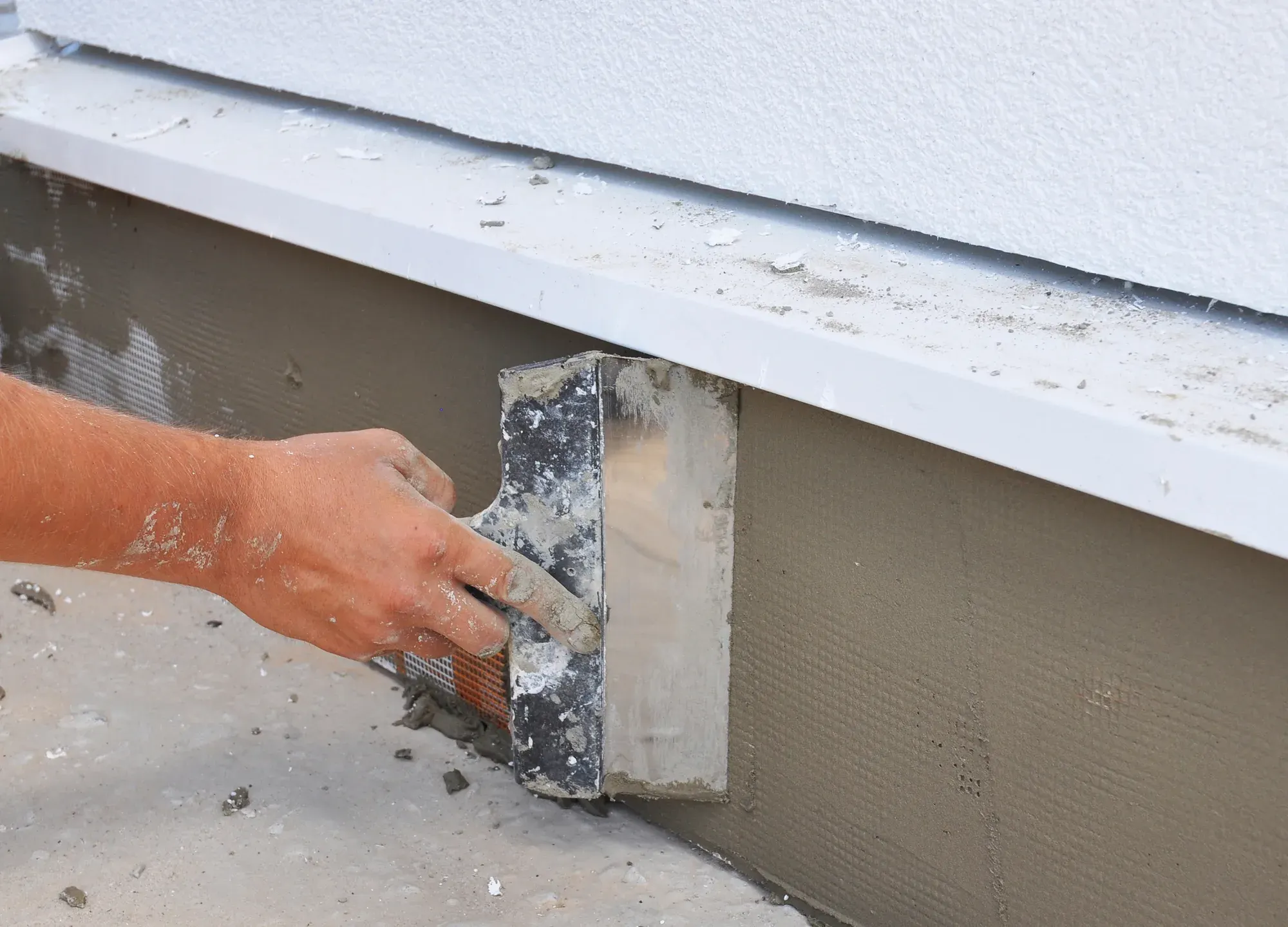 Trowel smoothing a fresh render coat along the base of a foundation